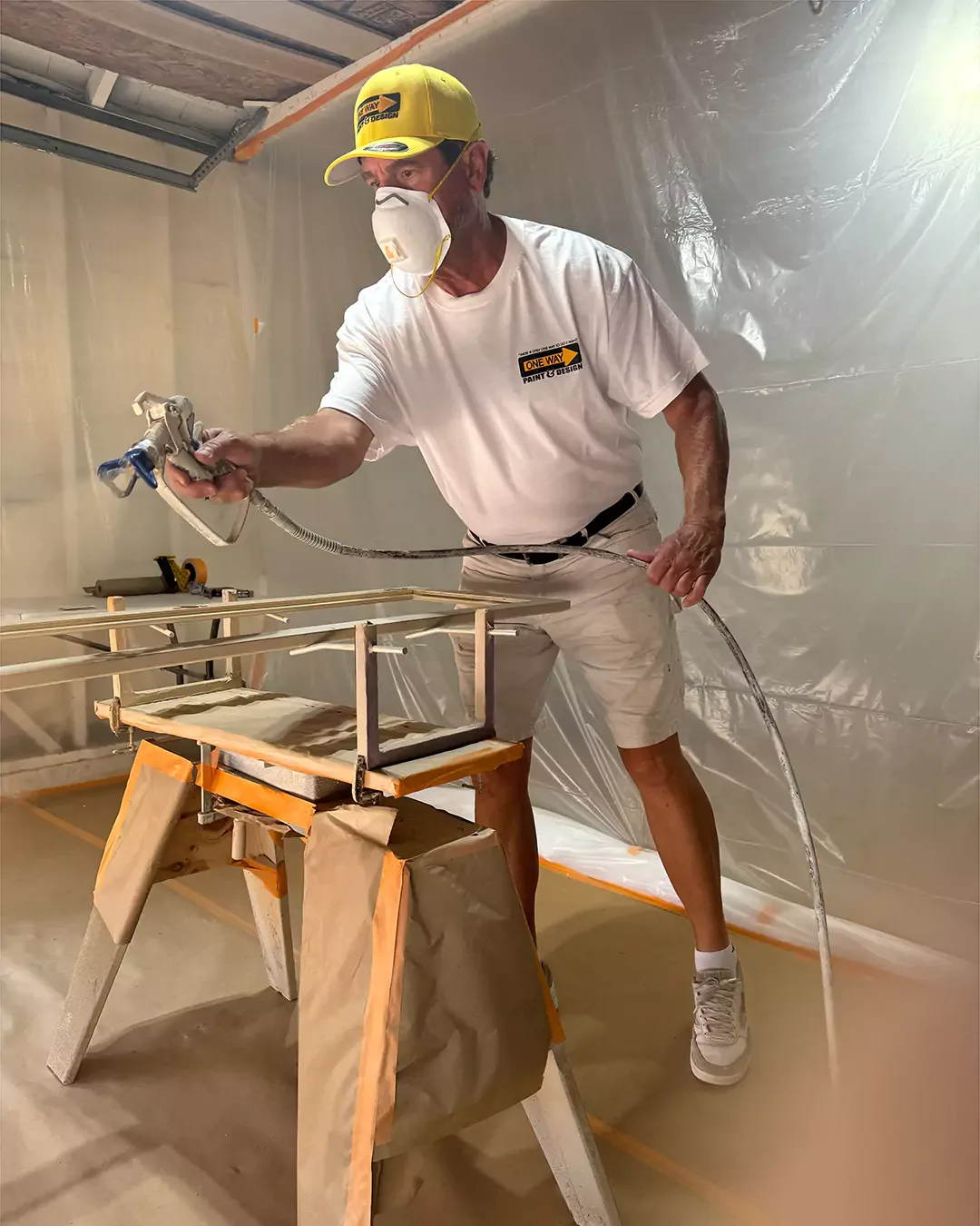 Danny Tron of One Way Paint & Design spray-finishing cabinet frames in a masked spray room, wearing the company's yellow cap